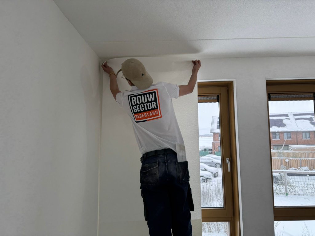 Behanger van Bouwsector Nederland zet een baan vlies tegen het plafond in een nieuwbouwkamer met besneeuwd uitzicht